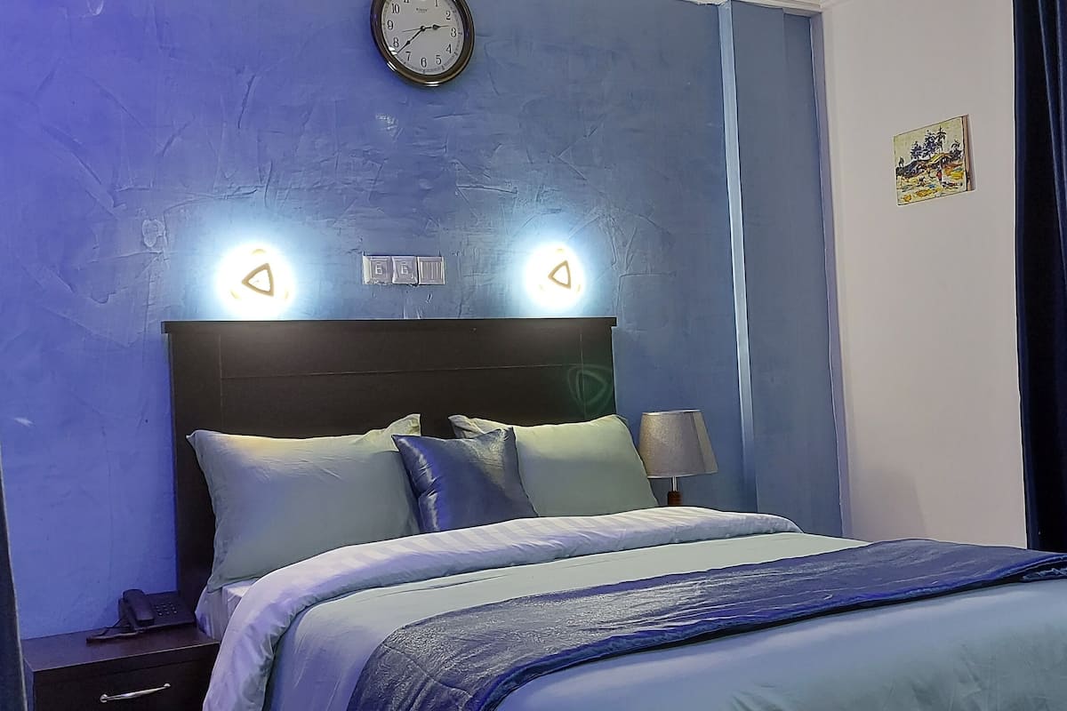 A neatly made bed with a headboard, two bedside lamps, and a clock on the wall.