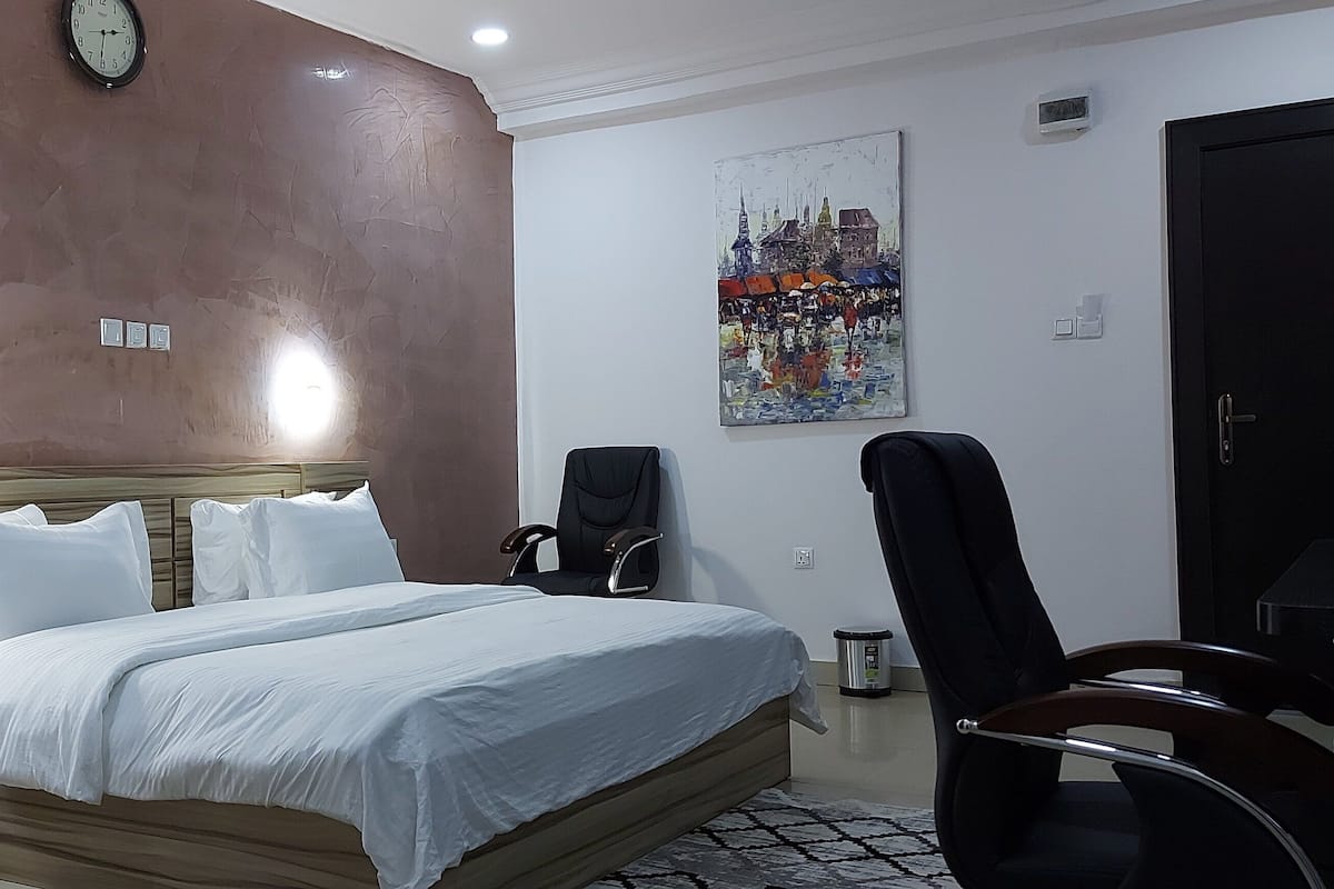 A hotel room with a bed, bedside lamp, clock, painting, and a door.