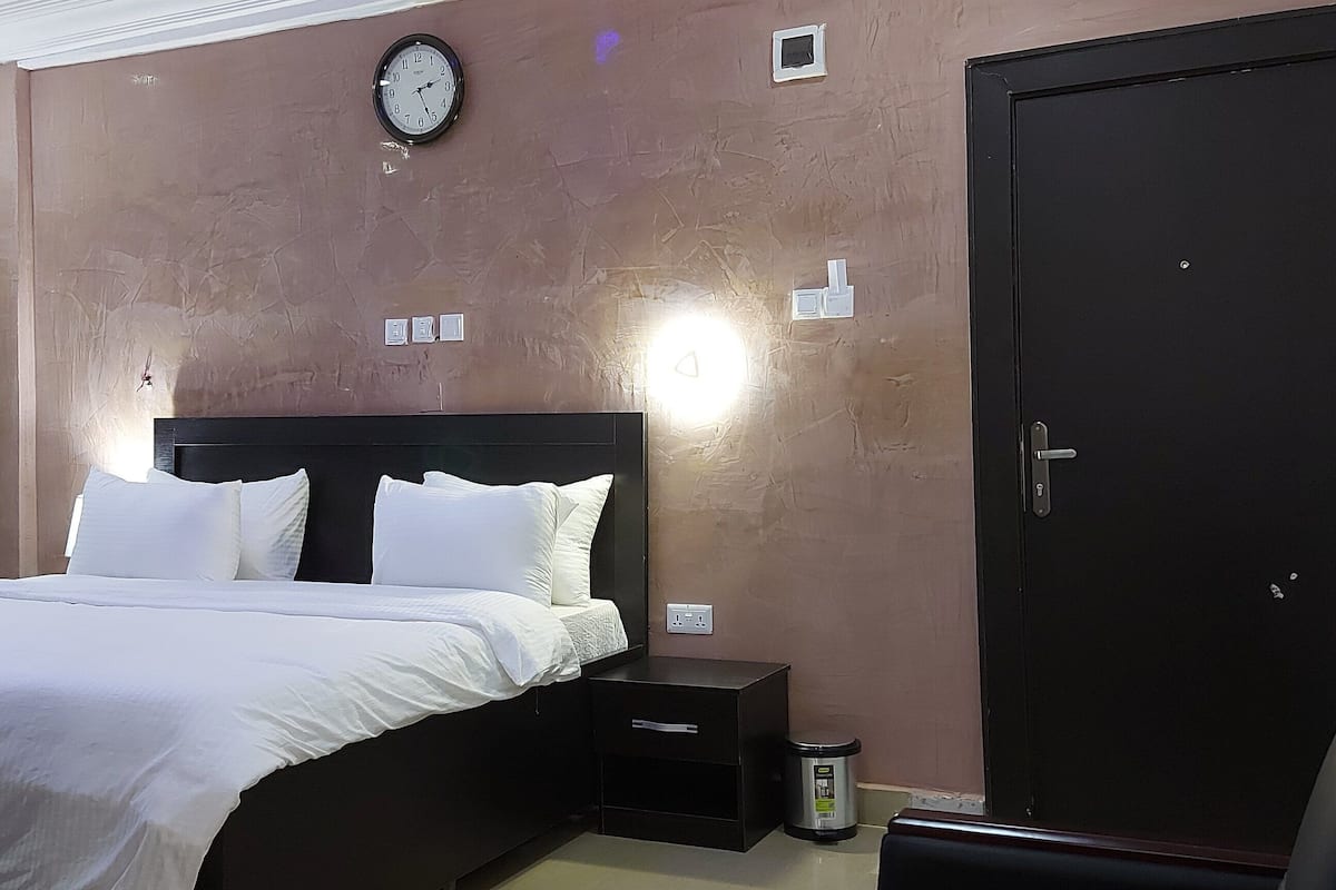 A hotel room with a bed, a nightstand, a trash bin, a wall clock, and a door.