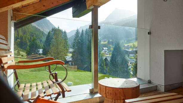 Een houten terras met een bankje en een ronde houten tafel, met uitzicht op een bergachtig landschap met huizen en bomen.