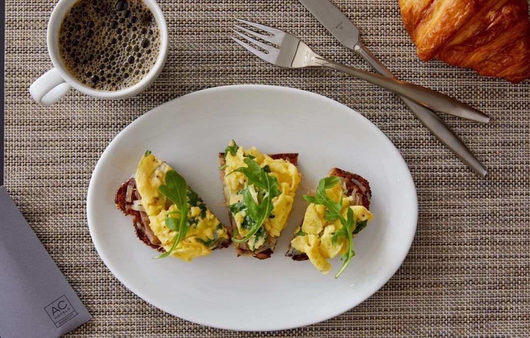 A plate with scrambled eggs on toast, a croissant, and a cup of coffee on a textured tablecloth.
