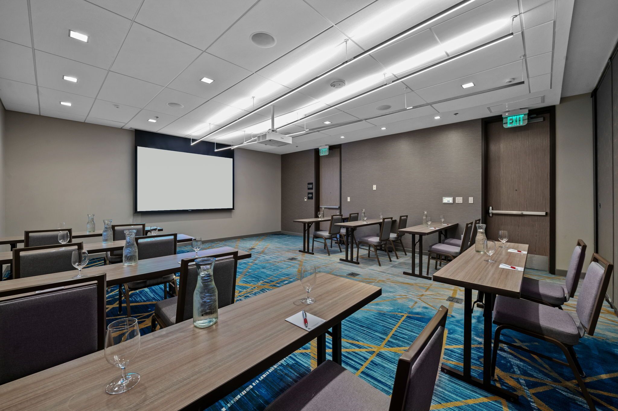 A conference room with tables, chairs, a projector screen, and a carpeted floor.