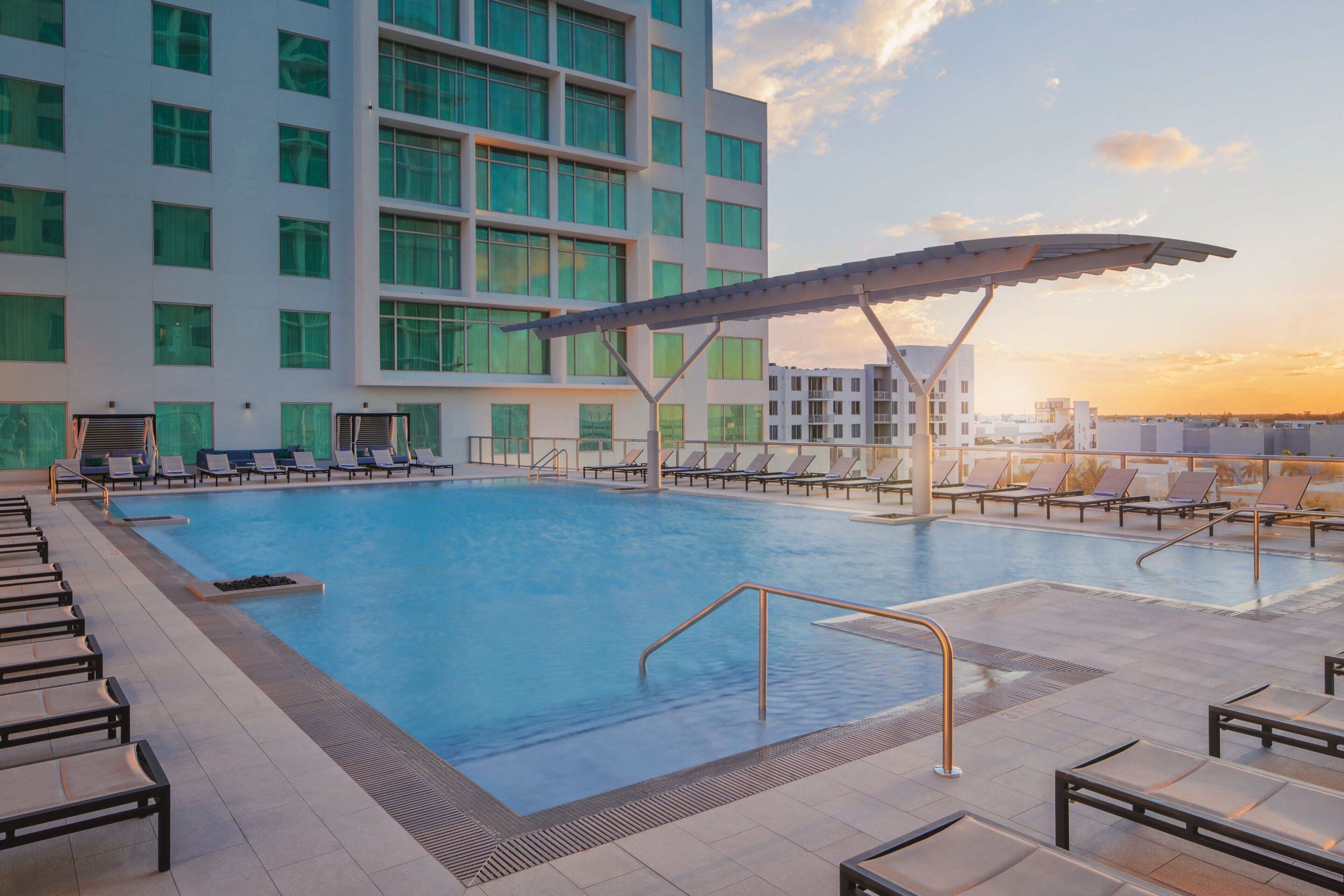 Piscina con sillas de descanso y una zona cubierta, rodeada por un edificio moderno con grandes ventanales.