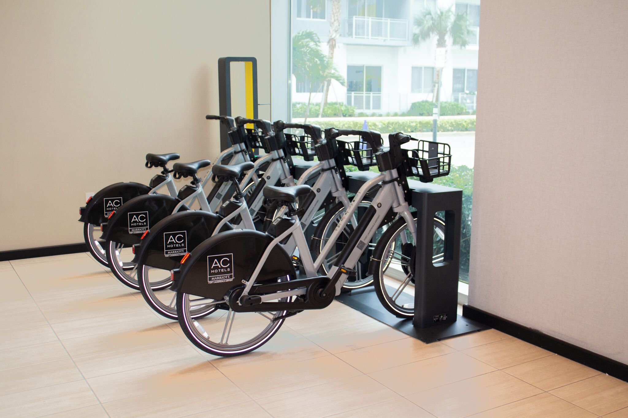 A row of stationary bikes with "AC Hotels" branding, located indoors with a view of palm trees and a building outside.