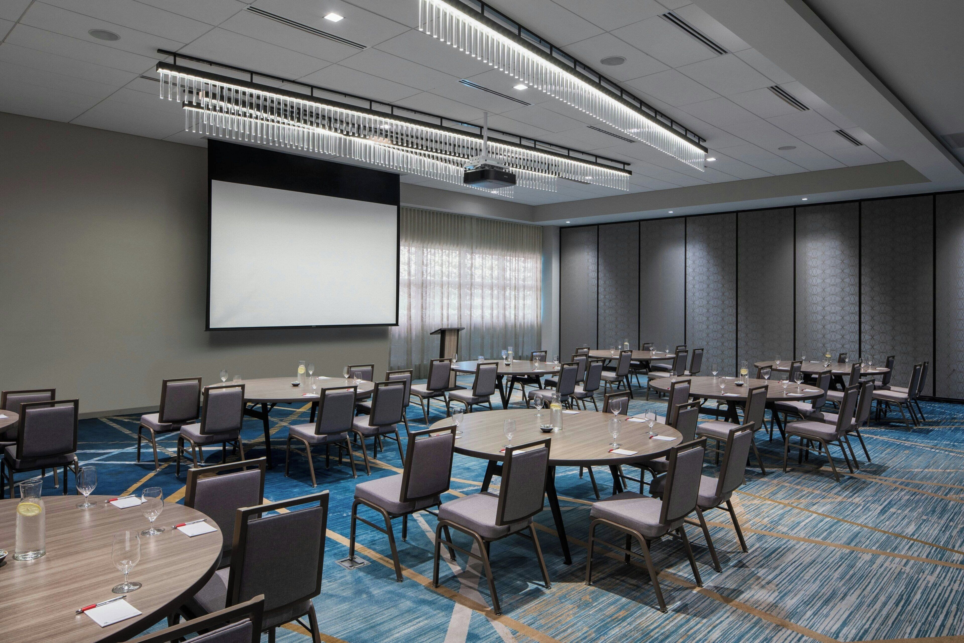 A conference room with multiple tables and chairs, a large screen, and a carpeted floor.
