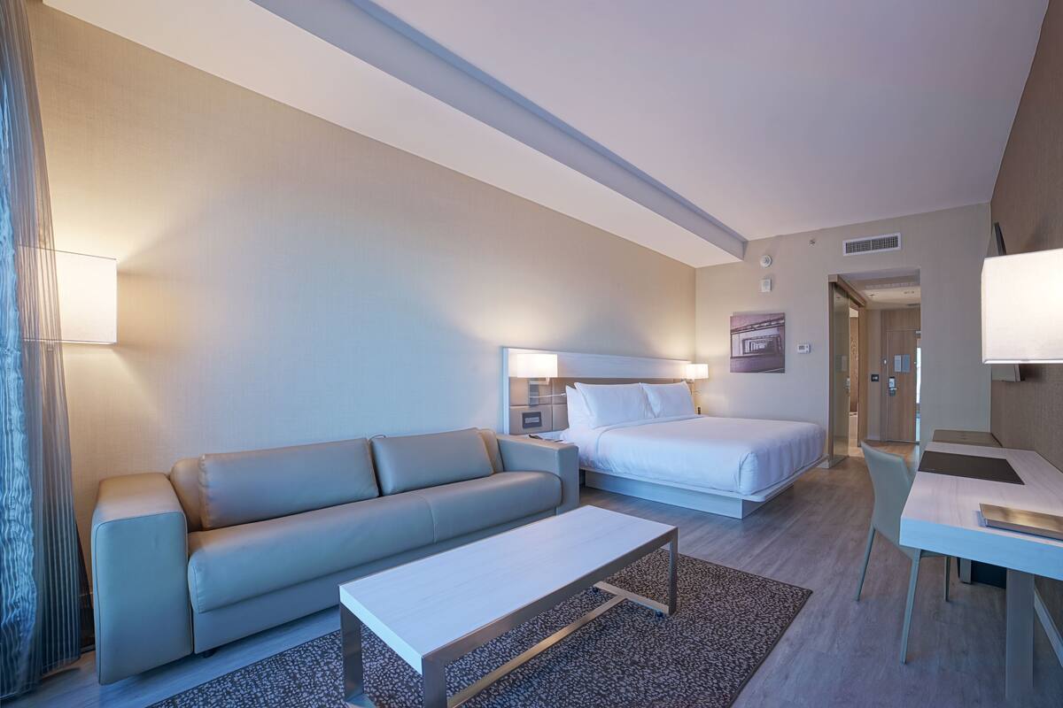 A modern hotel room with a sofa, a bed, a desk, and a lamp.