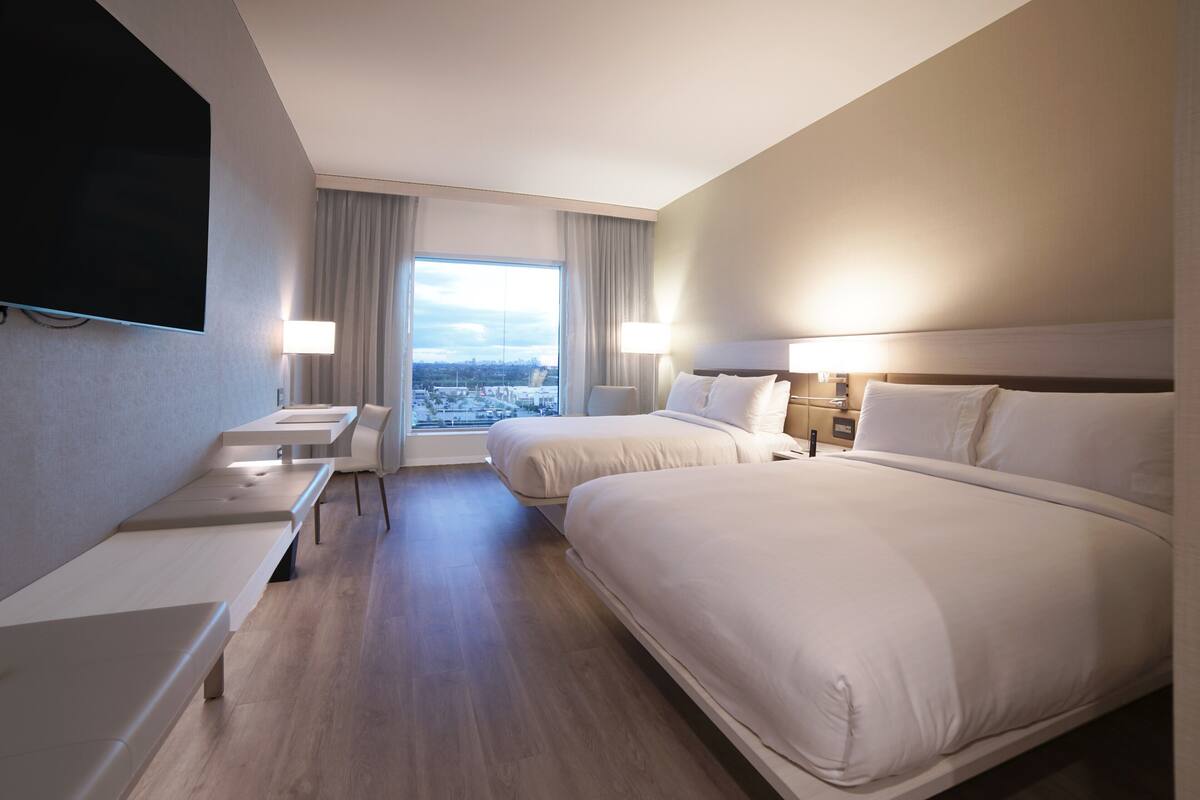 A hotel room with two beds, a TV, a desk, and a large window offering a city view.