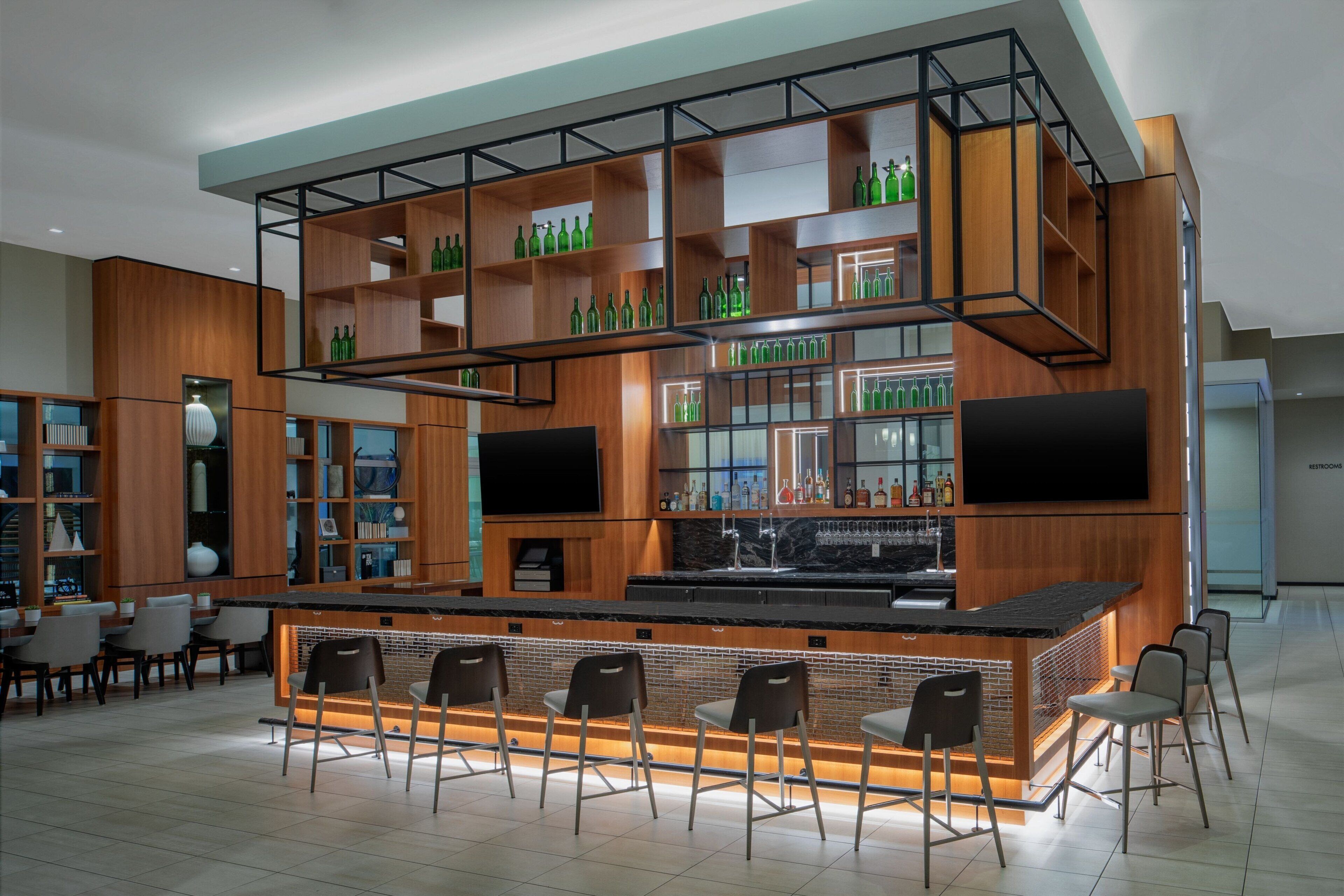 Un área de bar moderna con una barra de madera, varias estanterías con botellas y un televisor de pantalla plana sobre la barra.