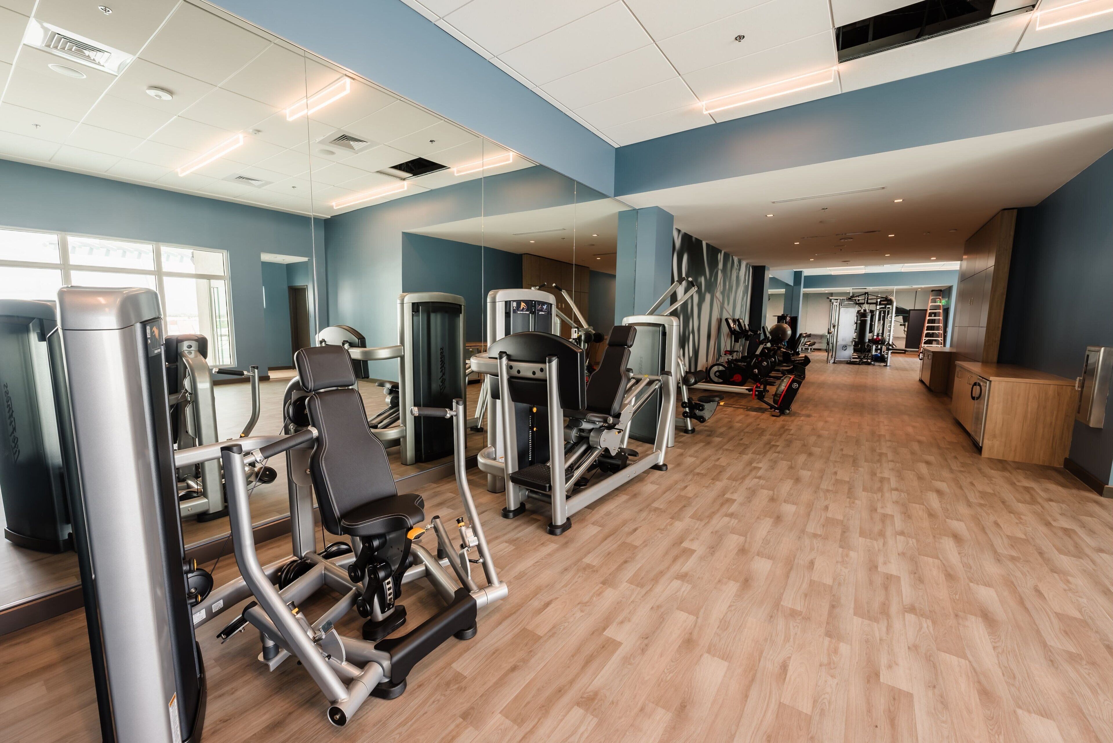 Un gimnasio con diversas máquinas de ejercicio, un espejo grande y suelo de madera.