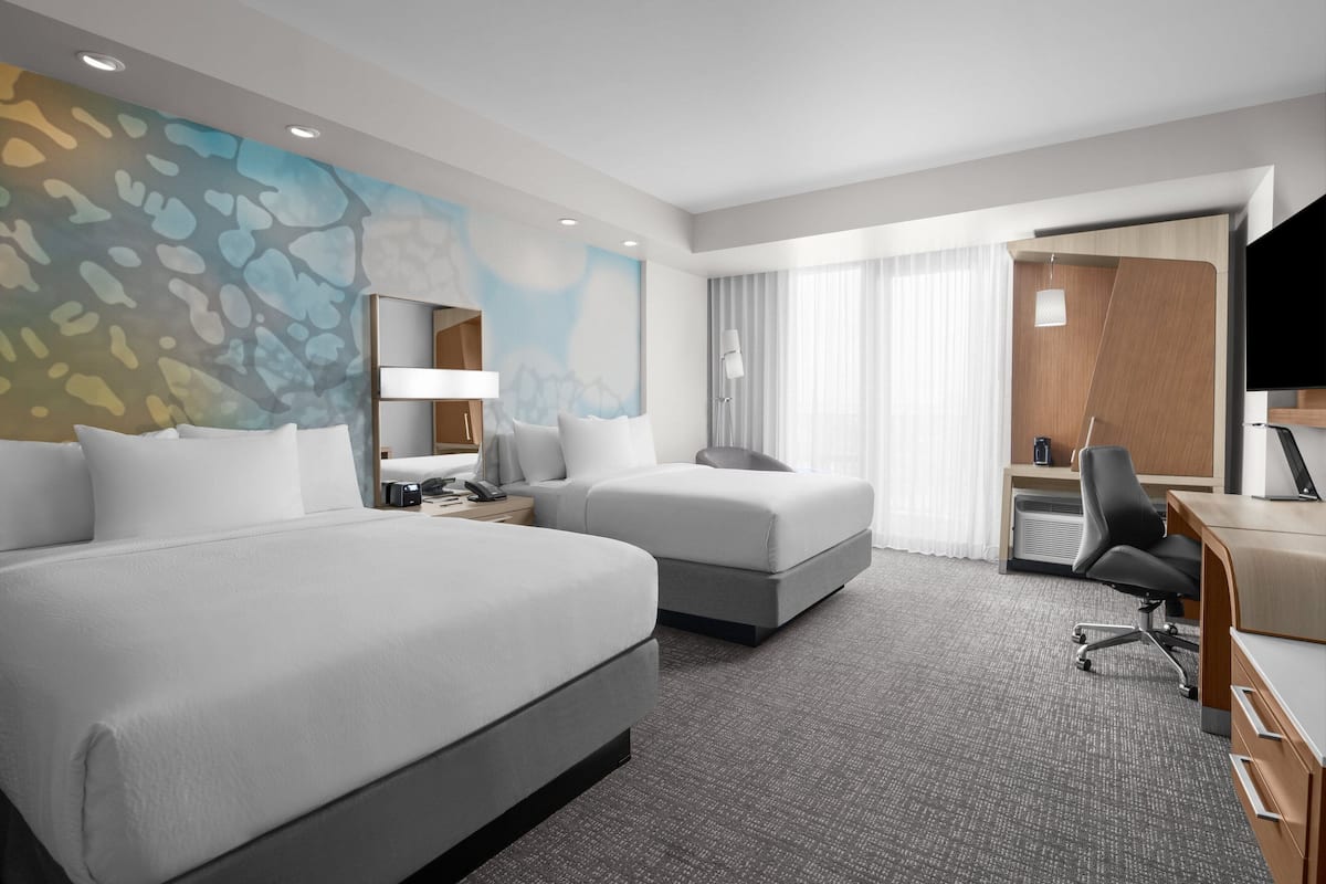 A hotel room with two beds, a desk, a chair, a TV, and a large abstract painting on the wall.