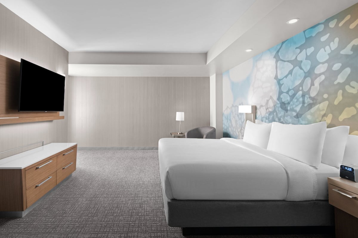 A modern hotel room with a large bed, a flat-screen TV mounted on the wall, a desk with drawers, and a nightstand with a lamp.