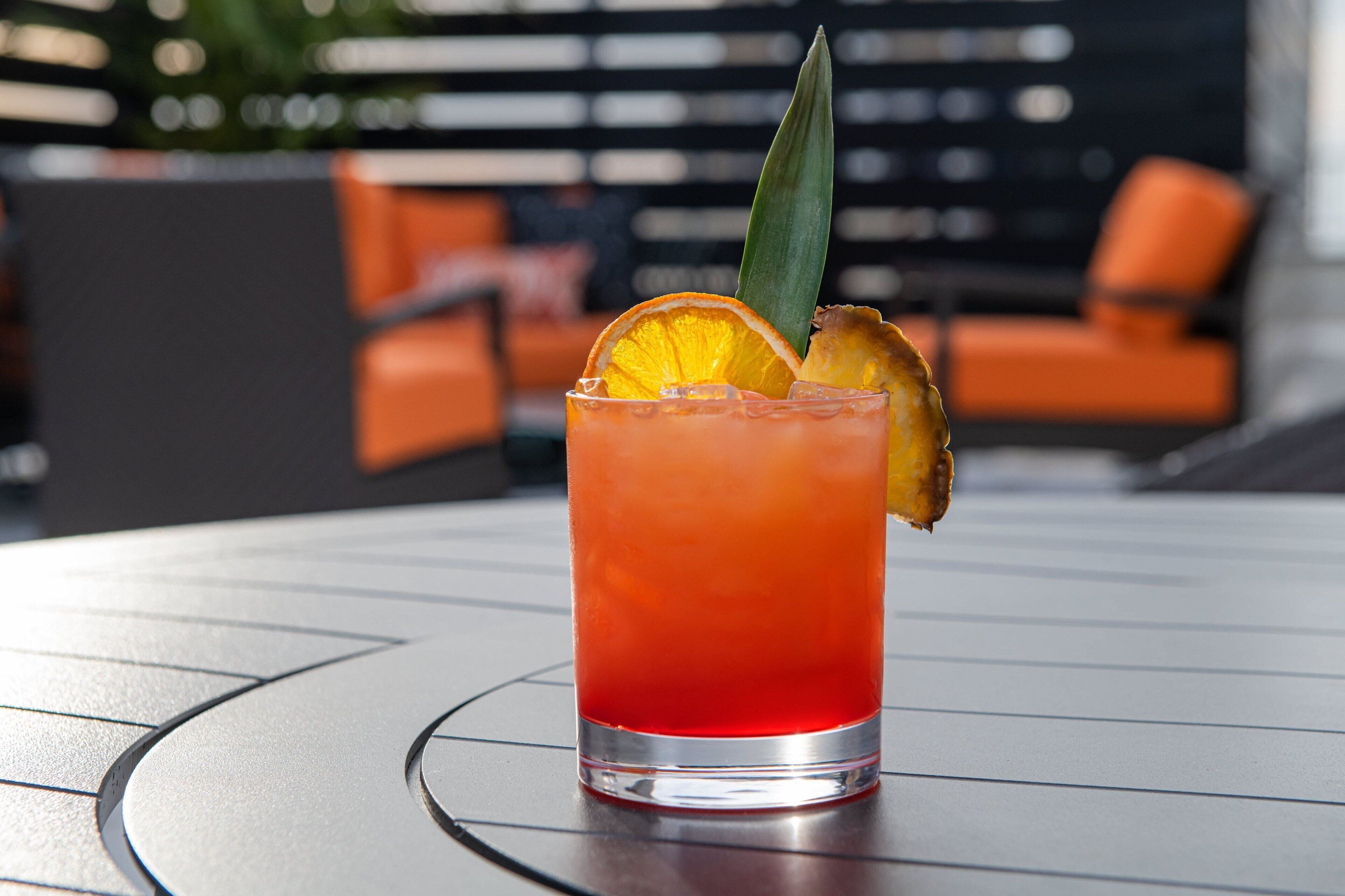 A glass of red drink with a slice of orange and a leaf garnish on a table.