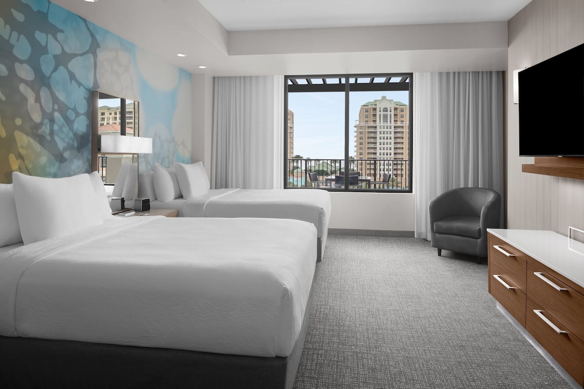 A hotel room with two beds, a TV, a desk, and a view of buildings.