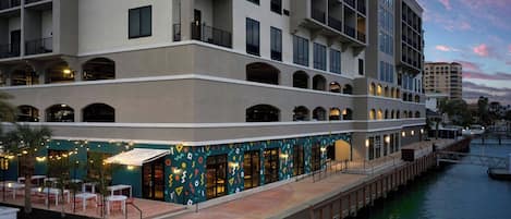 A waterfront hotel with multiple balconies, outdoor seating, and a covered walkway along the water.