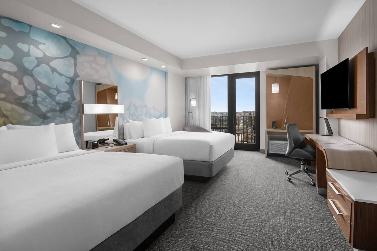 A hotel room with two beds, a desk, a TV, and a balcony with a view.