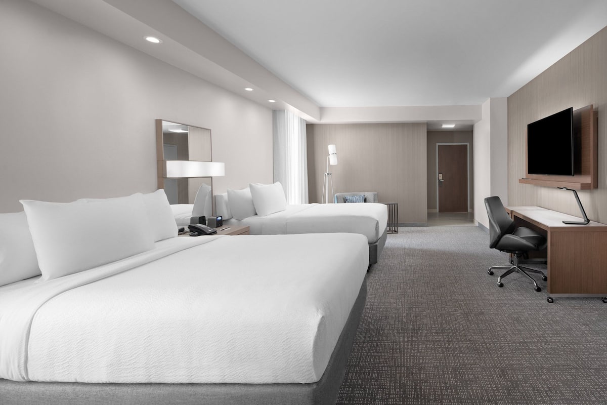 A hotel room with two beds, a desk, a chair, a TV, and a lamp.