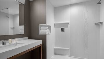 A modern bathroom with a large mirror, a white sink, a wooden vanity, a shower with a rain showerhead, and a towel rack.