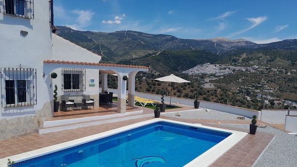 Ein Swimmingpool mit klarem, blauem Design, eine Terrasse mit Gartenmöbeln und Blick auf eine gebirgige Landschaft.