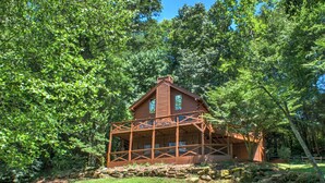 A rustic log cabin with a porch and stone foundation, surrounded by dense green trees. - Hot Tub, Pool Table & Fireplace + Biltmore Pass | Pet-Friendly Mountain Stay (Waynesville)