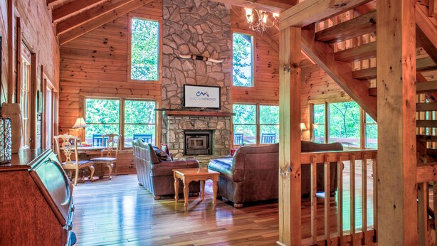 A spacious cabin interior with a stone fireplace, wooden floors, and large windows offering views of the outdoors. - Hot Tub, Pool Table & Fireplace + Biltmore Pass | Pet-Friendly Mountain Stay (Waynesville)