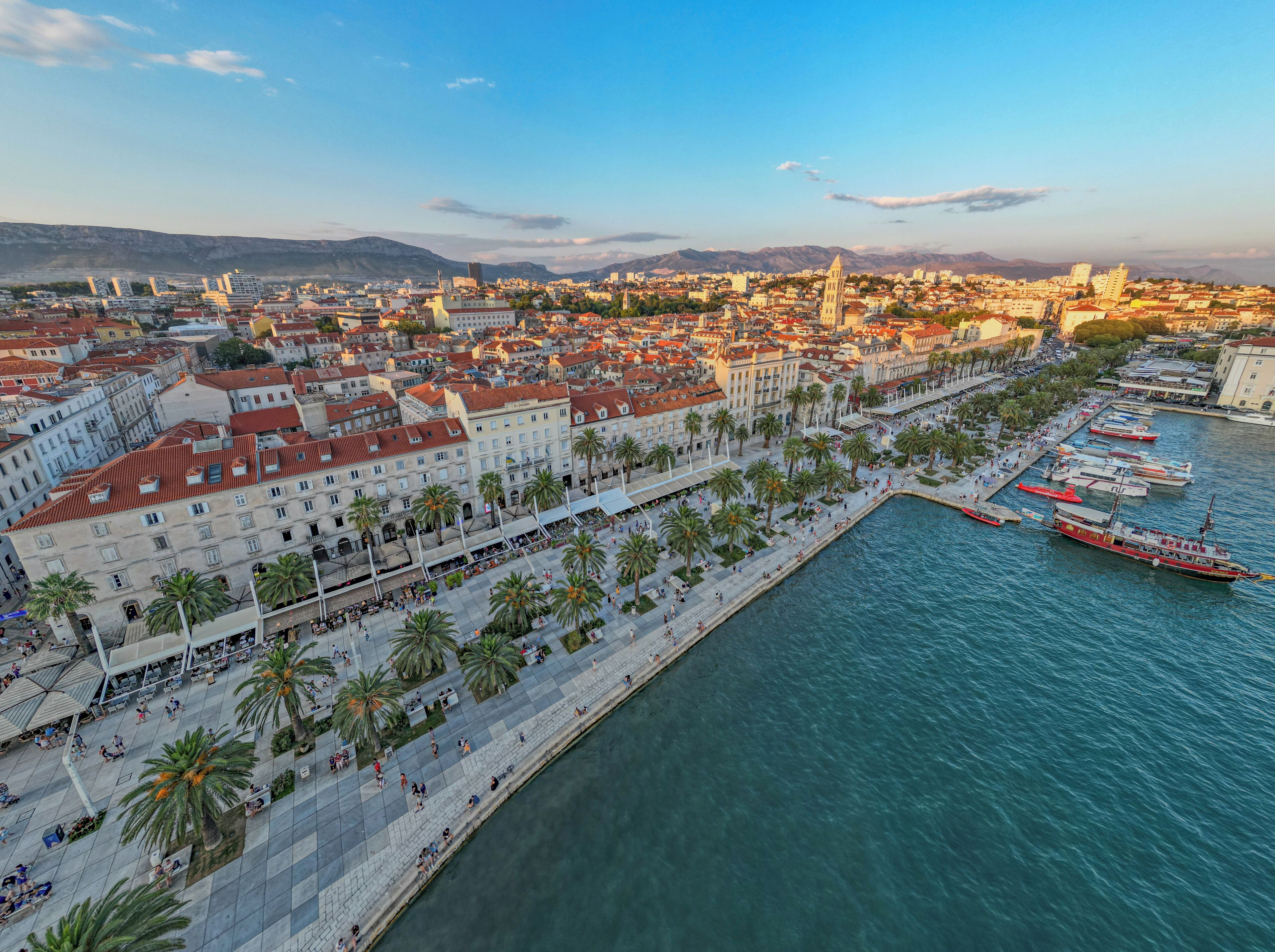 A coastal cityscape with a prominent waterfront promenade lined with palm trees and historic buildings.