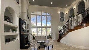 A spacious living room with a fireplace, a large window, a staircase, and a chandelier. - Florida Modern Mediterranean Villa (Cape Coral)