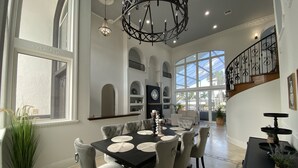 A spacious dining area with a black table and grey upholstered chairs, a chandelier, and a view of the outdoors through large windows. - Florida Modern Mediterranean Villa (Cape Coral)