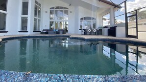 A swimming pool with a tiled edge, a covered patio area with seating, and large windows. - Florida Modern Mediterranean Villa (Cape Coral)