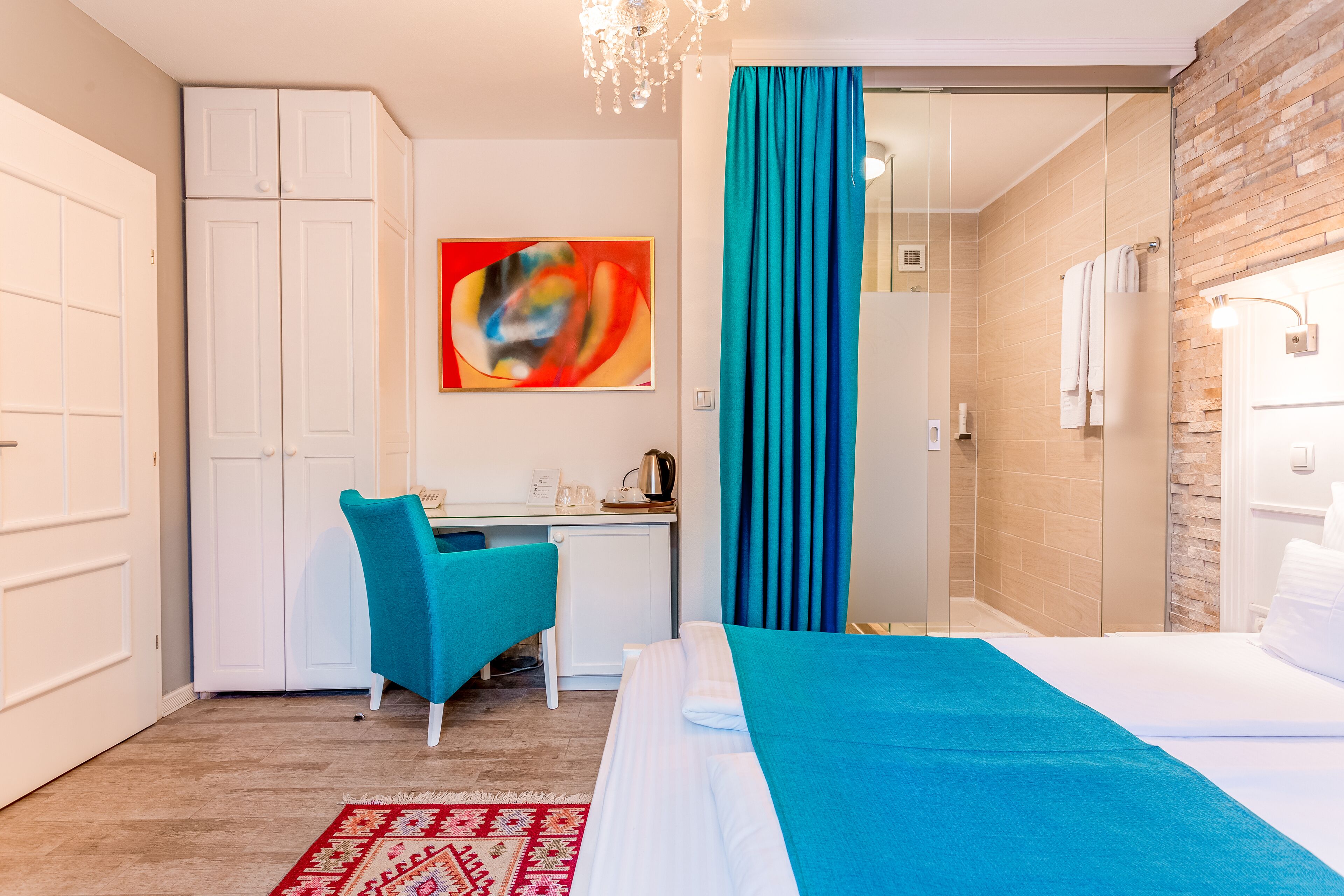 A hotel room with a bed, a desk, a chair, a red patterned rug, and a large abstract painting on the wall.