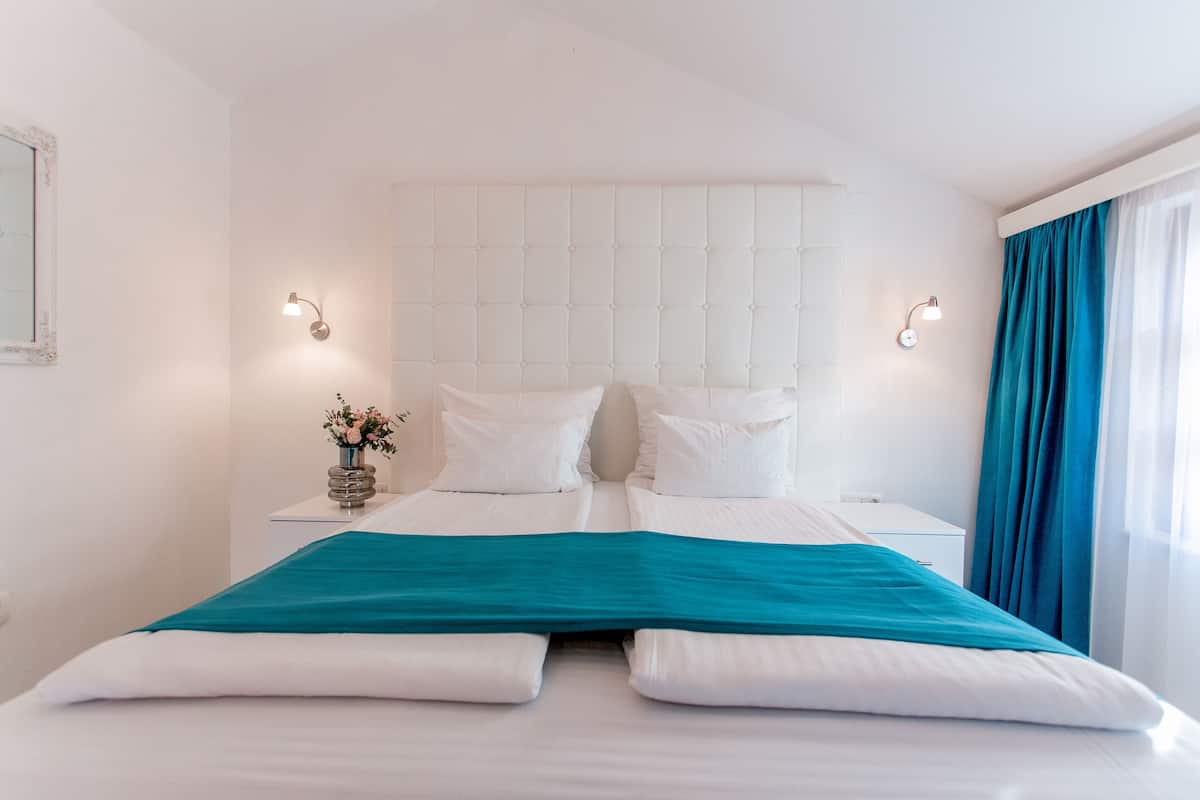 Un lit bien fait, avec du linge de lit blanc et un détail turquoise, une table de chevet ornée d’un vase de fleurs et une fenêtre avec des rideaux bleus.