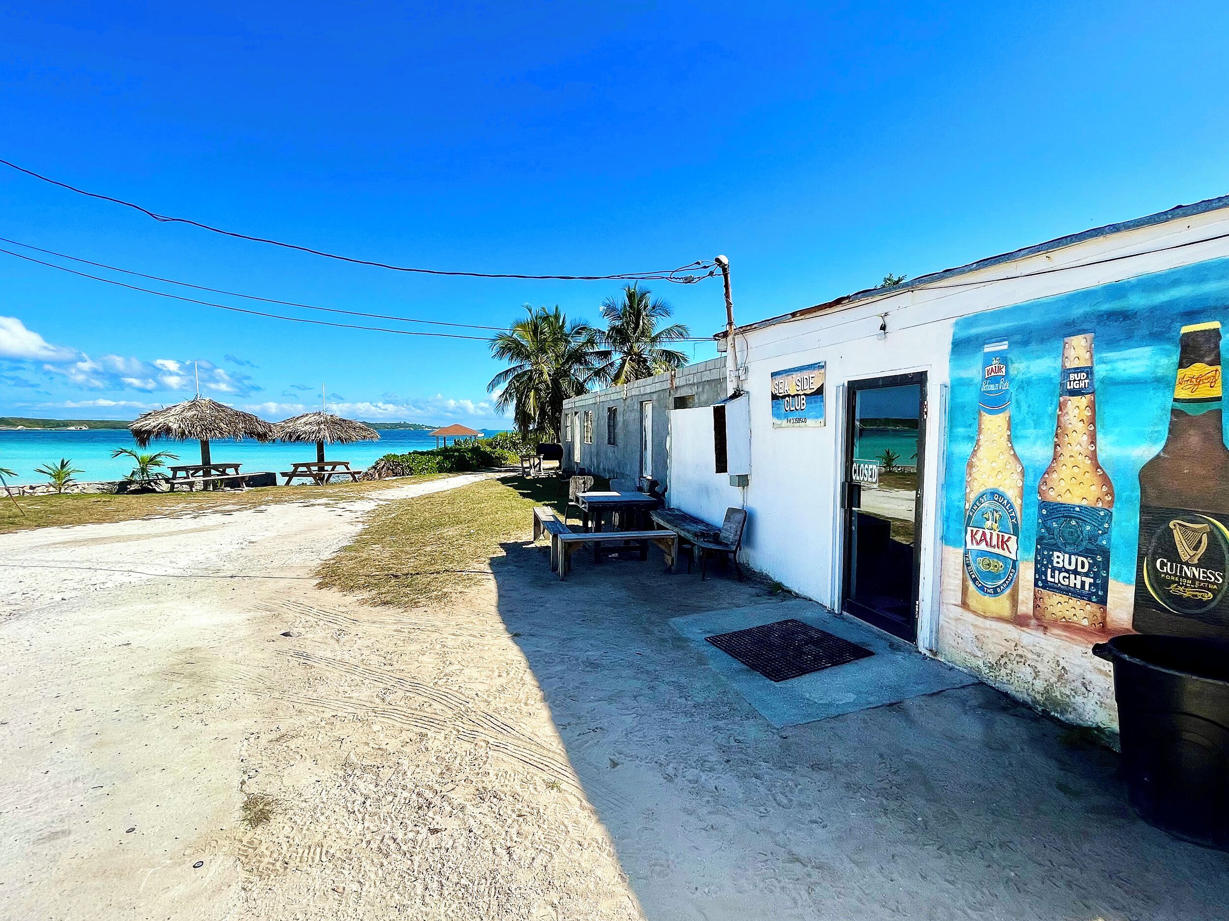 En bygning ved stranden med et mural af ølflasker, et skilt der siger 'SEA SIDE CLUB', og udendørs siddepladser ved vandet.
