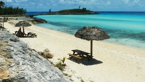 Una spiaggia con acque turchesi cristalline, una riva sabbiosa e una piccola isola all'orizzonte. Lungo la spiaggia si trovano panchine di legno e ombrelloni di paglia.