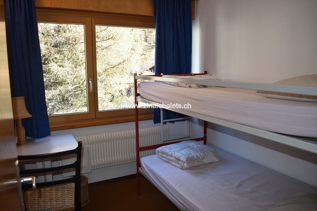 A bunk bed room with a view of trees outside, a desk with a lamp, and a chair.