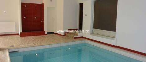 A swimming pool with a tiled floor and walls, a red-tiled shower area, and a large window.