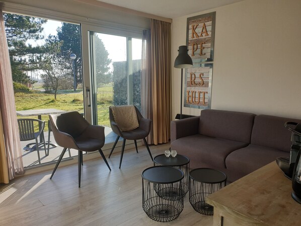 A modern living room with a sofa, two armchairs, a coffee table, and a small round table. There is a large window with a view of a garden and trees.