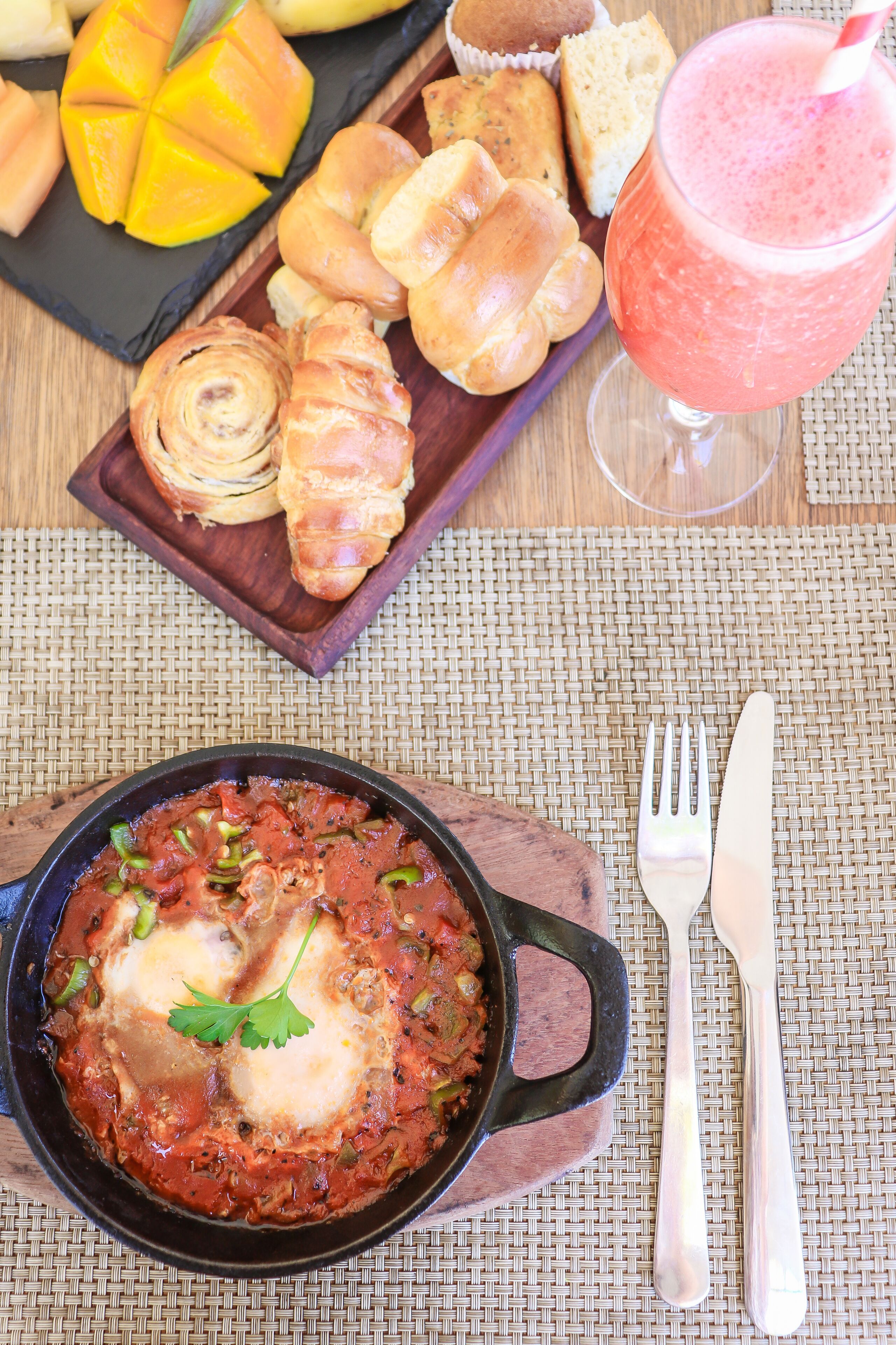 A meal with a bowl of shakshuka, a plate of pastries, a glass of smoothie, and a basket of fruit.