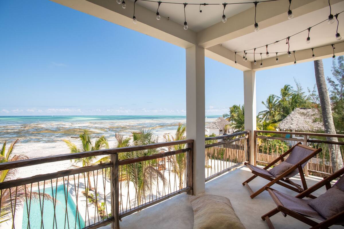 A balcony with a view of the beach and ocean, wooden lounge chairs, and string lights.