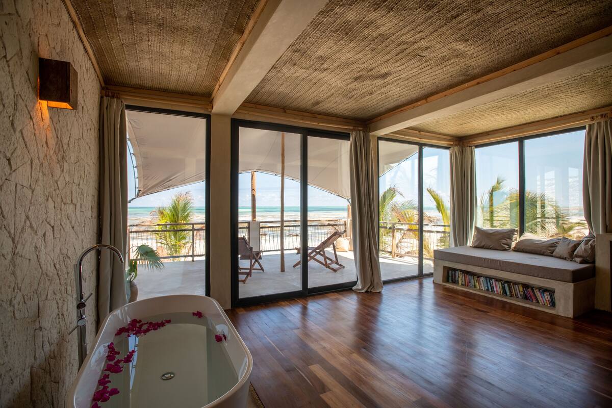 A spacious room with a large bathtub, a sofa with a built-in bookshelf, and a balcony with a view of the beach.
