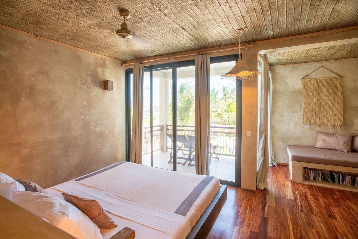 A bedroom with a large bed, wooden flooring, a balcony with a table and chairs, and a hanging light fixture.