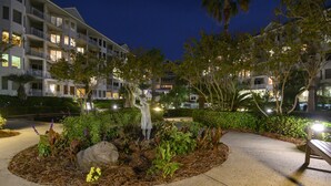 A landscaped garden area with a statue, surrounded by well-maintained bushes and trees, in front of a multi-story hotel with balconies and illuminated windows. - 2303 Seacrest - 2 Oceanfront Pools, Beach Bar, & Steps to the Beach! (Hilton Head Island)