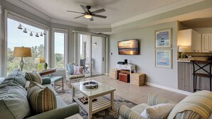 A spacious living room with a large sofa, a coffee table, and a television mounted on the wall. There are framed pictures on the wall, a ceiling fan, and a view of the outdoors through large windows. - 2303 Seacrest - 2 Oceanfront Pools, Beach Bar, & Steps to the Beach! (Hilton Head Island)