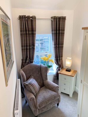 Une pièce chaleureuse avec une fenêtre ornée de rideaux à carreaux, un fauteuil moelleux, une table de chevet avec une lampe et un radiateur.