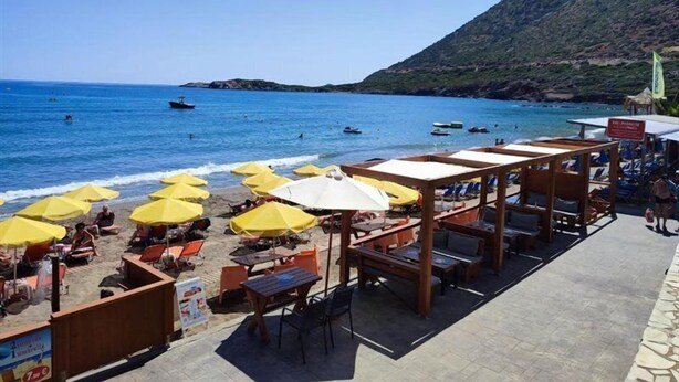 Un café frente a la playa con terraza y sombrillas, con vista a un mar tranquilo con barcos.