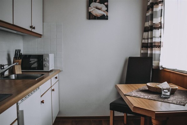 Una cocina con armarios blancos, un microondas y un lavabo. Hay una mesa de comedor con una silla y una cesta.