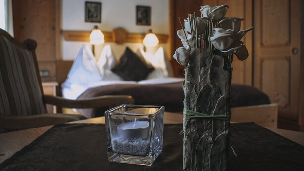 A hotel room with a bed, a bedside table with a candle and a vase of flowers, and wooden walls. - Landhaus Andrea (Saalbach-Hinterglemm)