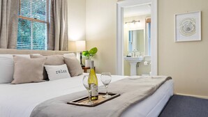 Habitación de hotel con cama, mesita de noche con una botella y vasos, espejo y ventana con cortinas.