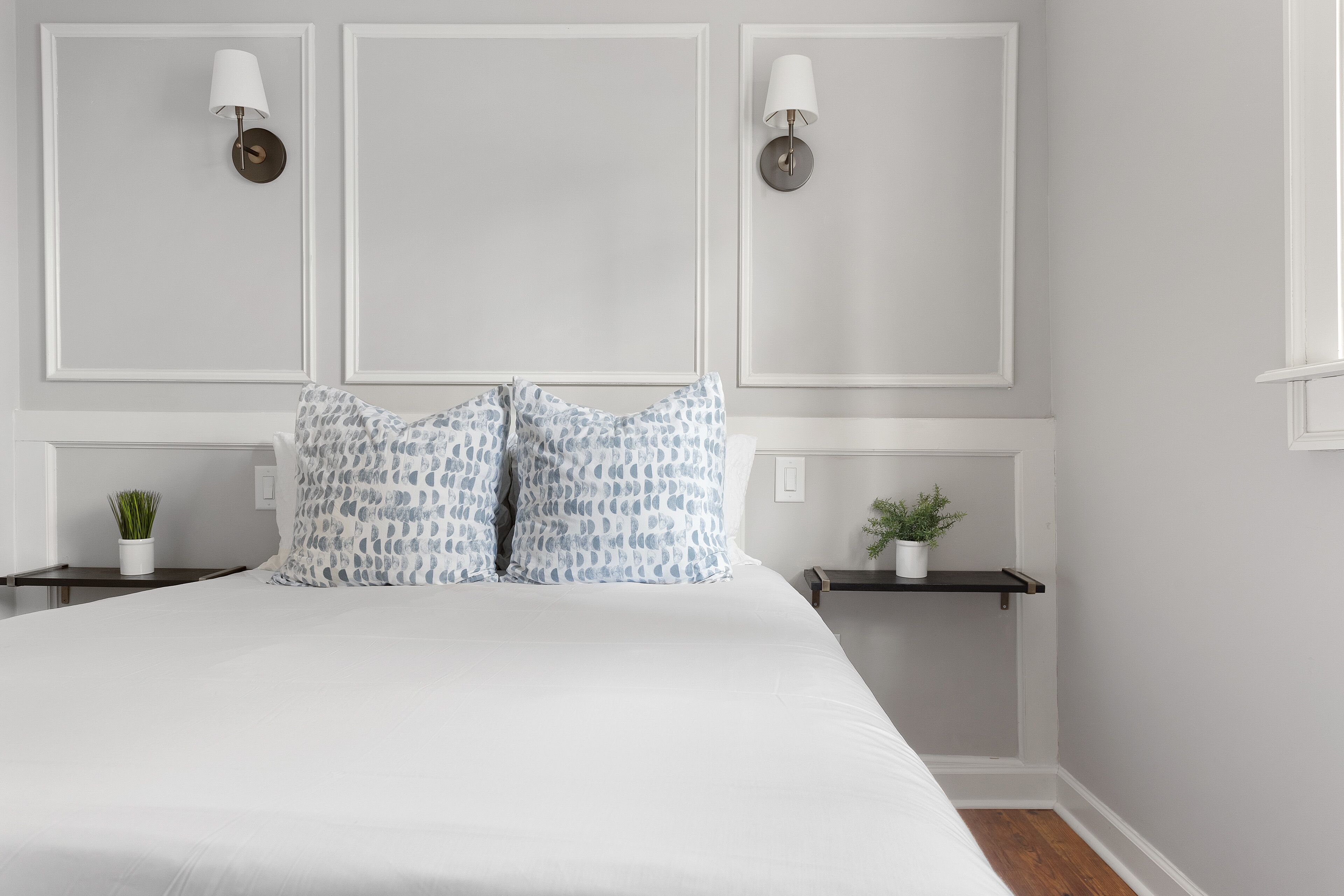 A neatly arranged bedroom with a large bed, two bedside tables with plants, and wall-mounted lamps.