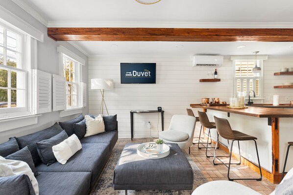 A modern living room with a grey sofa, a coffee table, and a bar area with stools. - Only 3 Blocks from King | Backyard! | The Felix (Charleston)