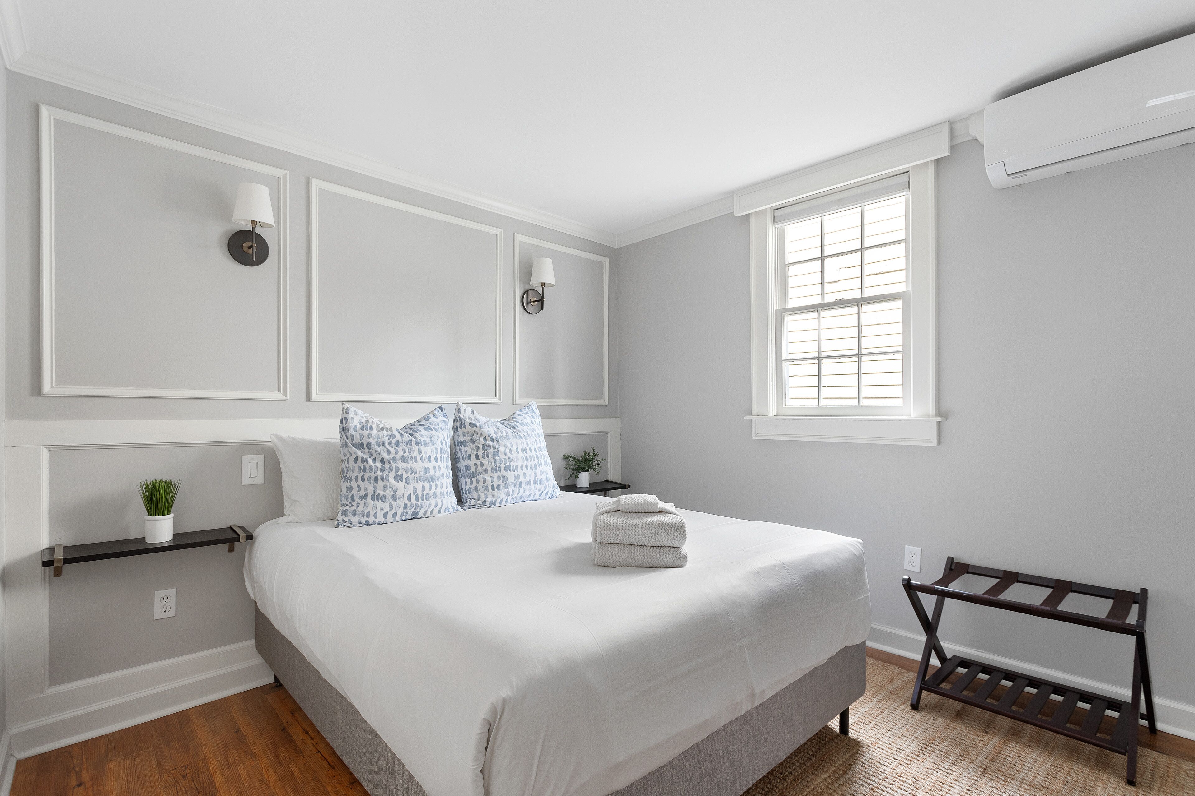 A bedroom with a large bed, two wall-mounted lamps, a window with blinds, and a small plant on a shelf.