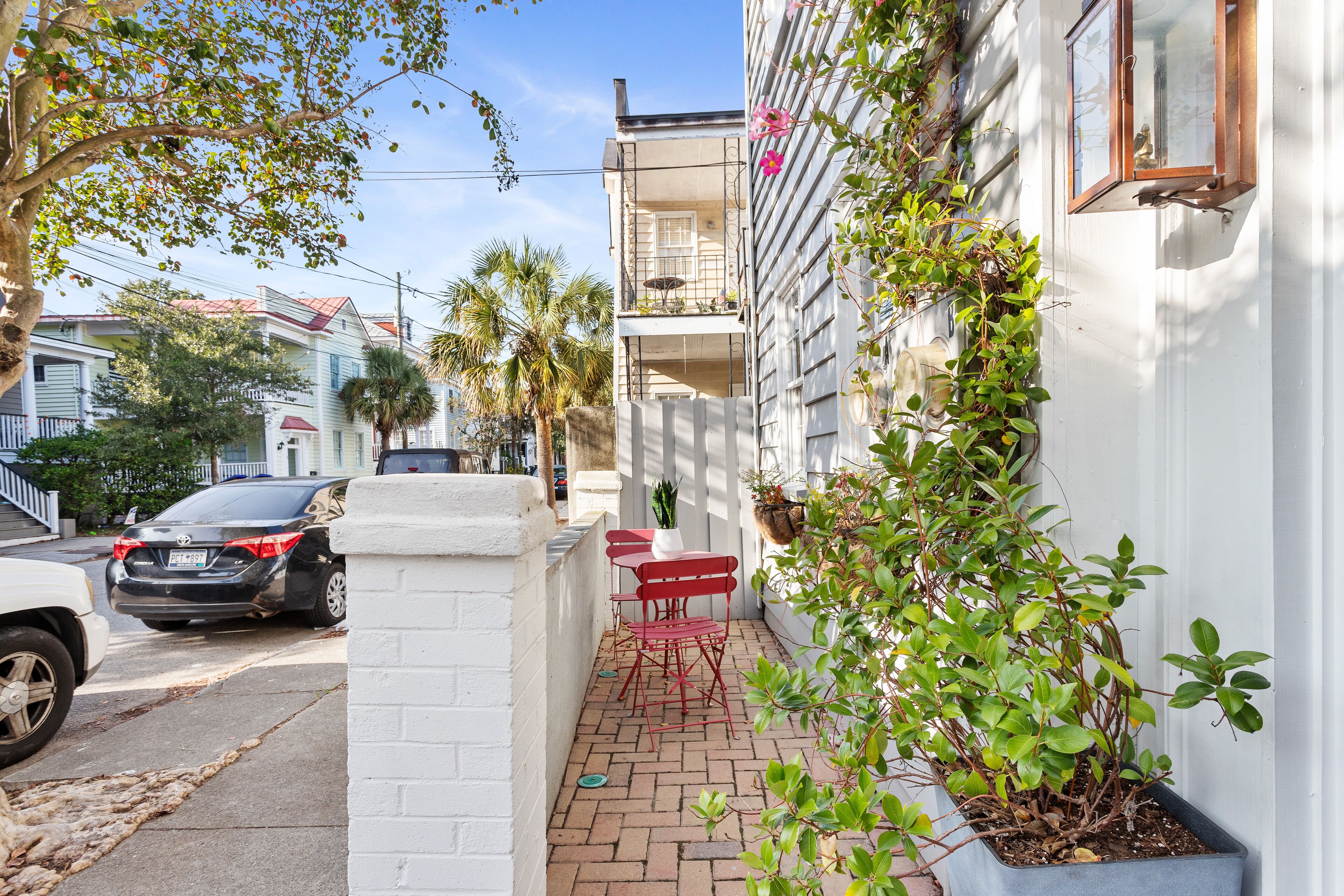 A narrow outdoor patio with red chairs and a table, adjacent to a white building with a balcony.